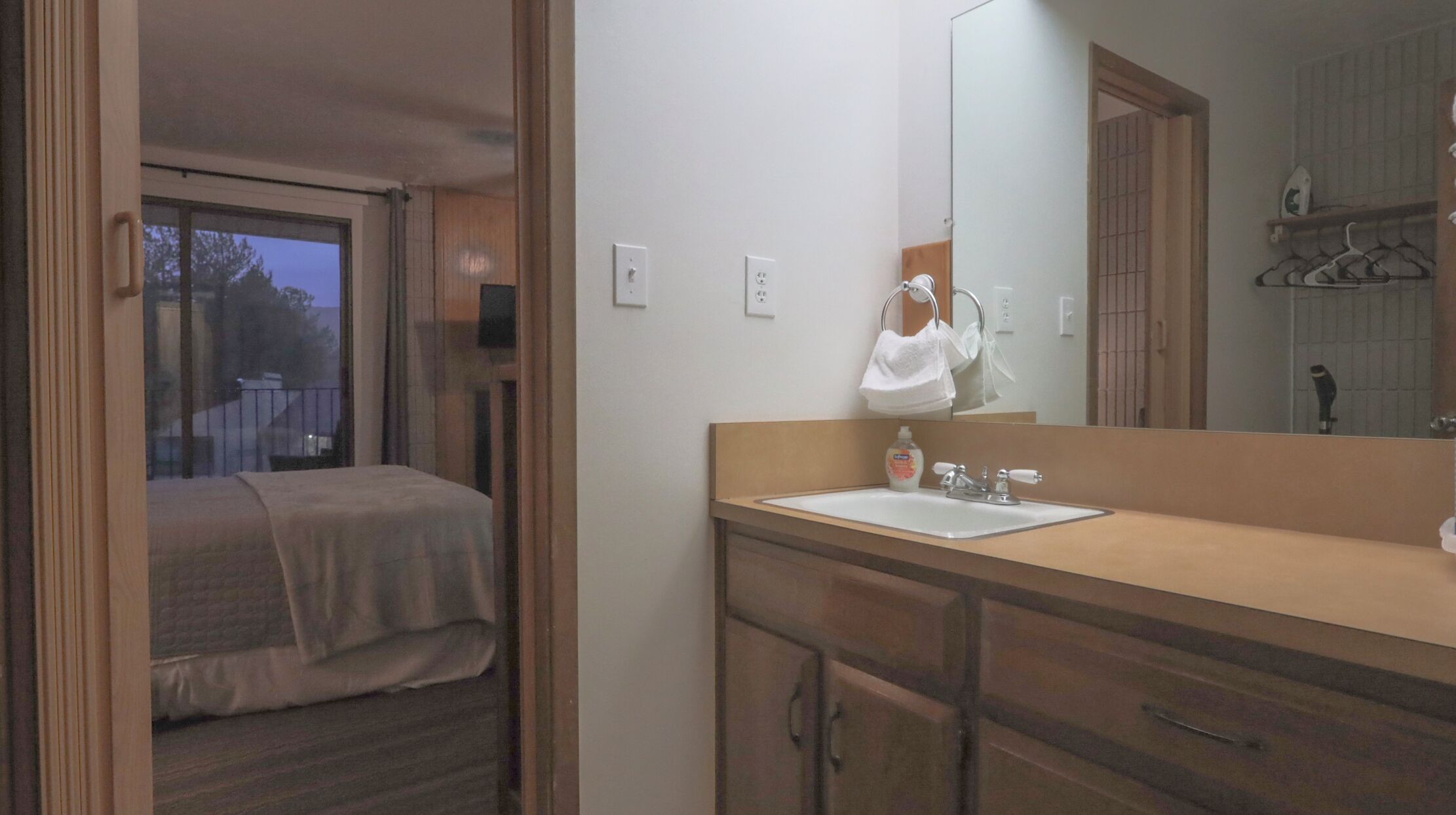A compact bedroom with a bed, a bathroom with a sink and mirror, and a view of the outside through a window.