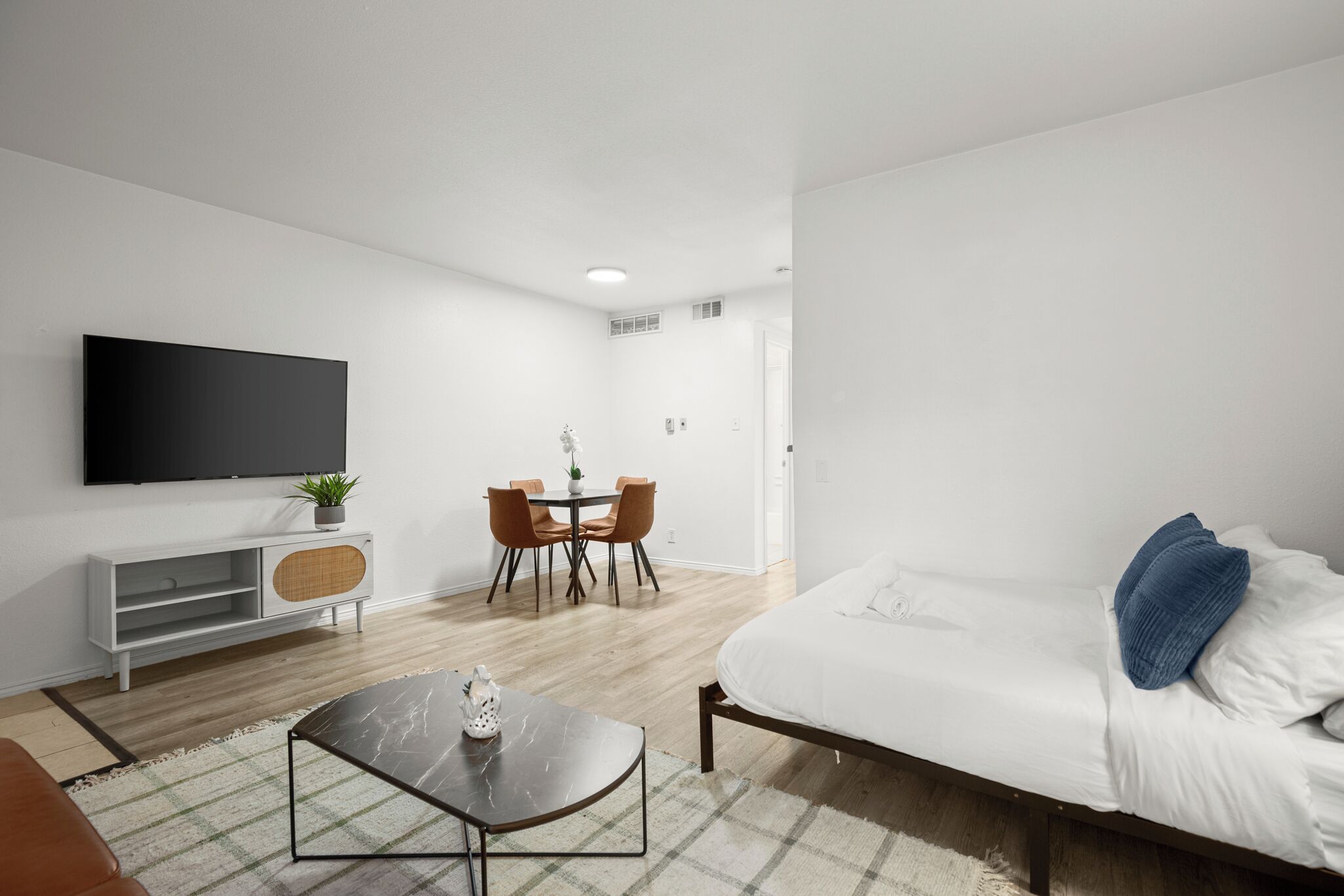 A modern, well-lit living room with a flat-screen TV, a dining area with a table and chairs, and a bed with a blue pillow.
