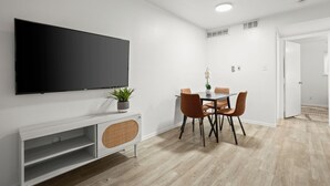 Ein modernes Wohnzimmer mit einem an der Wand befestigten Flachbildfernseher, einem Esstisch mit vier Stühlen, einer kleinen Kommode mit einer Pflanze und einer Tür zu einem anderen Raum.