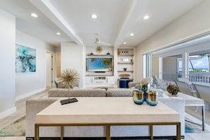 Ein modernes Wohnzimmer mit einem großen Sofa, einem Couchtisch und dekorativen Accessoires. Ein Fernseher steht auf einem Schrank und ein Fenster bietet Blick aufs Meer.