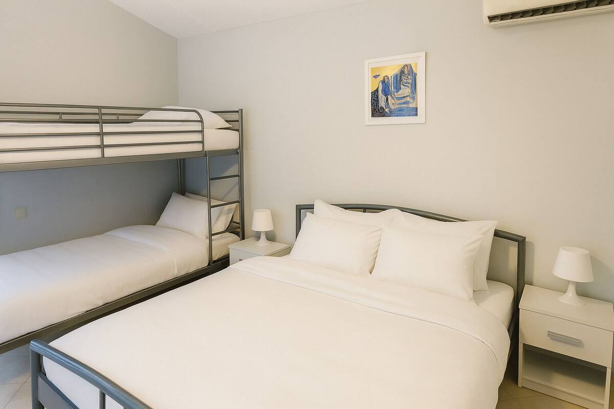 Een hotelkamer met stapelbedden, een enkele bed, nachtkastjes met lampen en een lijst met een kunstwerk aan de muur.
