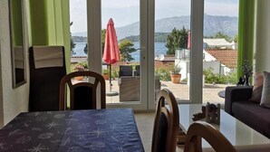 A room with a table and chairs, a sofa, and a view of a lake and mountains through large sliding glass doors. - Lumbarda Resort Apartments (Lumbarda)