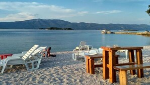A beach with white plastic chairs and a wooden table, a calm sea, and mountains in the background. - Lumbarda Resort Apartments (Lumbarda)