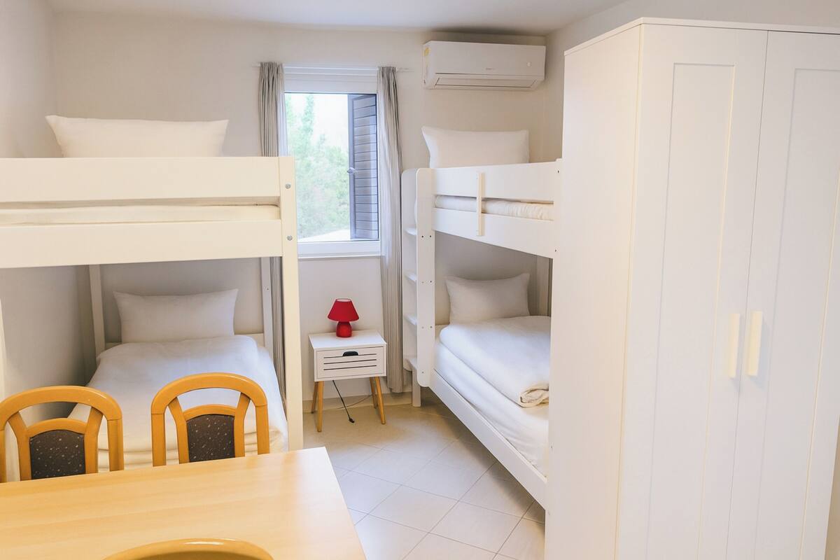 Een slaapkamer met stapelbedden, een kleine tafel met stoelen, een nachtkastje met lamp en een kledingkast.