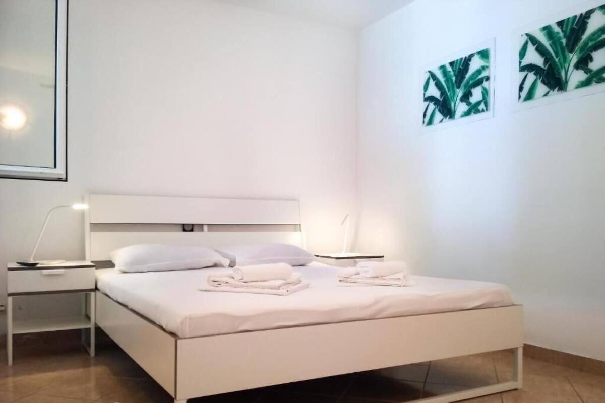 Een opgemaakt bed met witte lakens en kussens, een nachtkastje met een lamp, en een muur met drie bladmotieven in lijsten.
