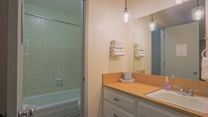 Een badkamer met een douche, een wastafel en een spiegel. Handdoeken hangen aan de muur en er is een zeepdispenser op de wastafel.