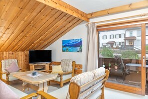 A cozy room with a sloped ceiling, wooden furniture, a flat-screen TV, and a large window offering a view of a building and greenery.
