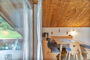 Un intérieur en bois avec une table et des bancs, une radio, et une vue sur un paysage verdoyant par la fenêtre.