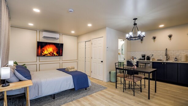 A modern bedroom with a bed, bedside table, and a TV displaying a fireplace. Adjacent is a compact kitchen with a dining area.