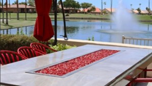 Une table à manger recouverte d’une nappe blanche, entourée de chaises rouges, le tout mis en valeur par un décor composé d’un bassin avec une fontaine, de palmiers et d’un ciel dégagé.