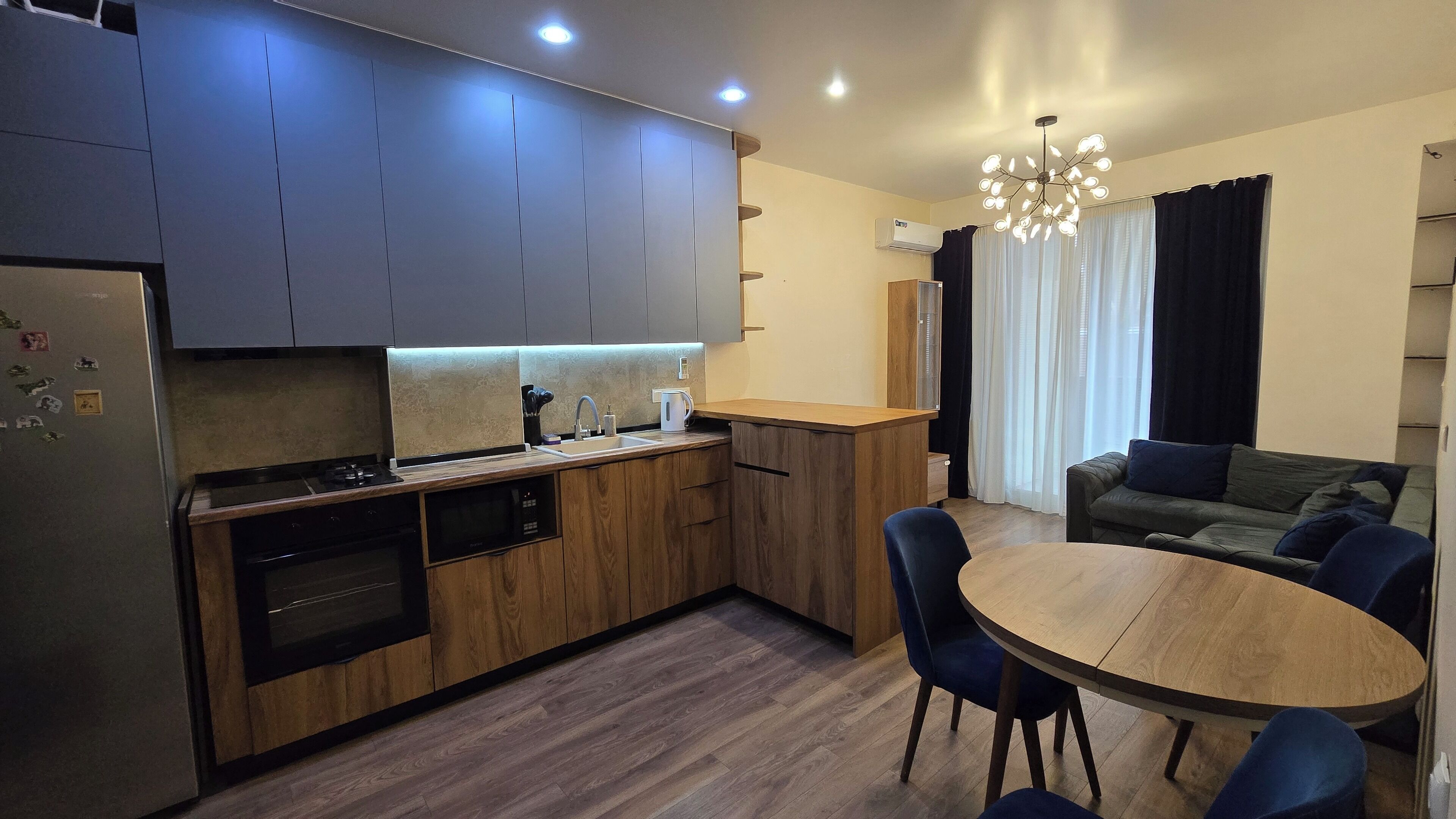 Eine moderne Küche mit Holzschränken, einem Einbauofen und einem Essbereich mit rundem Tisch und blauen Stühlen.