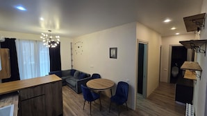 Ett modernt vardagsrum med en soffa, ett runt bord och blå stolar. Här finns också ett matbord i trä och en köksdel med en diskho och skåp.