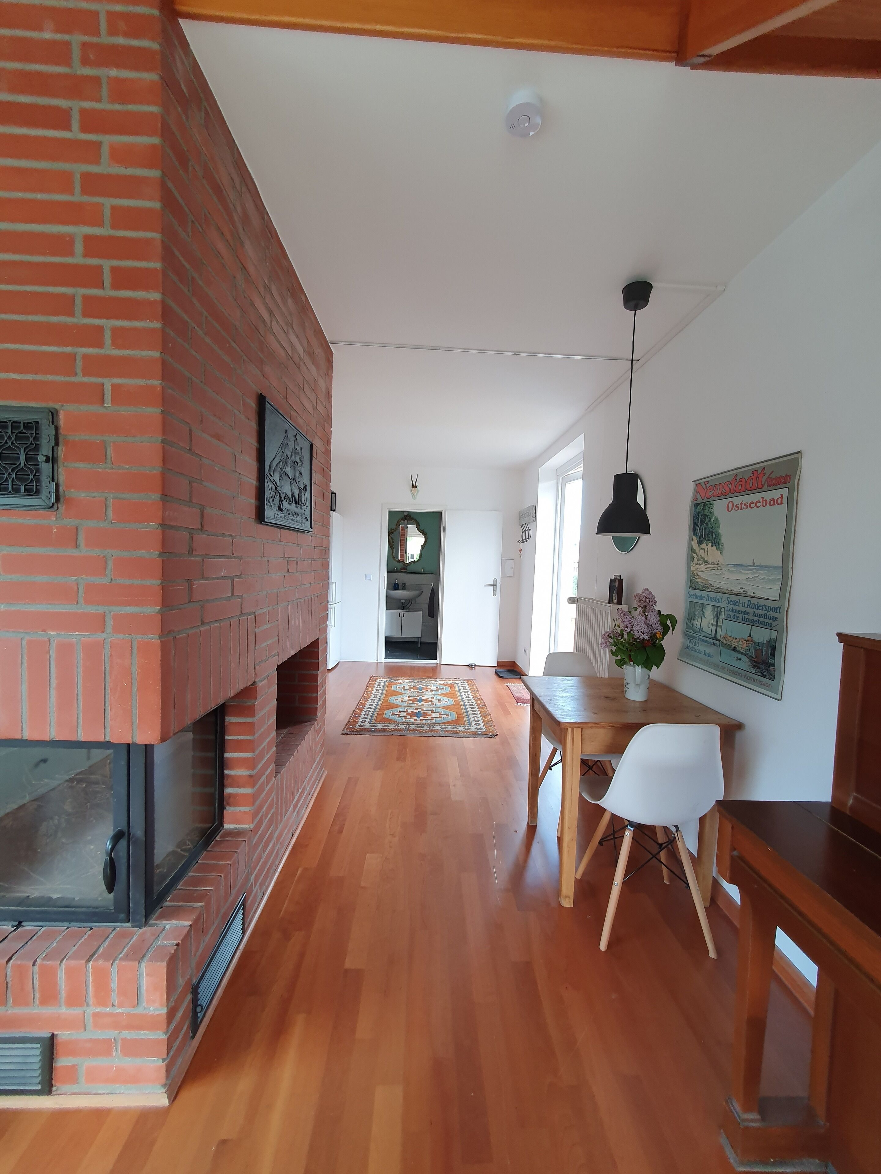 Ein geräumiger Raum mit Kamin aus Ziegelsteinen, Holzboden, einem Esstisch mit Stühlen und einer hängenden Pendelleuchte.