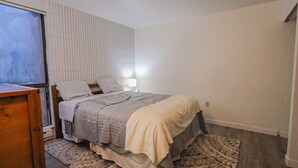 A bedroom with a bed, a nightstand, a lamp, a window with blinds, and a patterned rug. - Quaint 1-bedroom condo - 112 Lava Hot Springs Resort Building A (Lava Hot Springs)