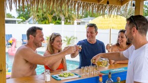 A bar area with a pool in the background, a thatched roof, and a wooden counter. - Backyard Oasis • Pool •  Hot Tub • Putt Putt • Bar (Largo)