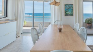 A modern dining area with a wooden table, white chairs, and a view of the sea through large windows. - Spectacular 2-Bedroom Apartment with Sea View – Market Square (LE LAVANDOU)