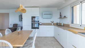 A modern kitchen with white cabinetry, a wooden dining table, and a hanging light fixture. - Spectacular 2-Bedroom Apartment with Sea View – Market Square (LE LAVANDOU)