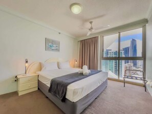Habitación de hotel con una cama grande, una mesita de noche, un ventilador de techo y vistas a la ciudad desde la ventana.
