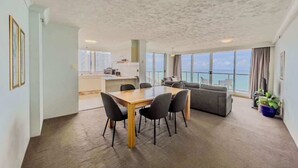 A spacious living area with a dining table and chairs, a sofa, and a large window offering a view of the sea. - Luxury 2 Bedroom Ocean View Apartment in Surfers Paradise (Surfers Paradise)