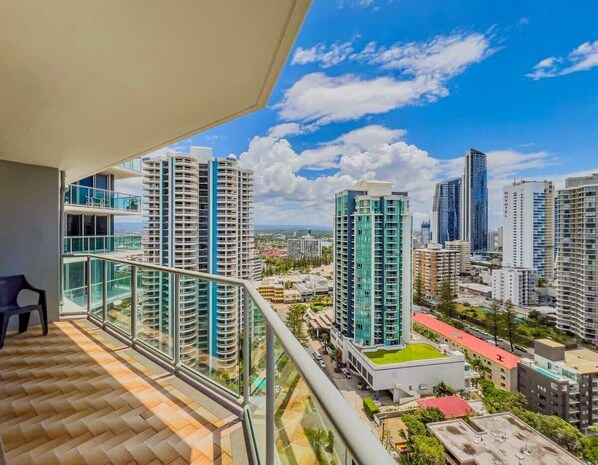 Un edificio alto con fachada de vidrio, un balcón con barandilla metálica y vistas a otros edificios y áreas verdes.