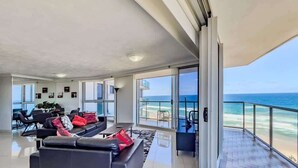 Ett rymligt vardagsrum med en stor soffa och fåtöljer, en balkong med utsikt över stranden och ett stort fönster med utsikt över havet.