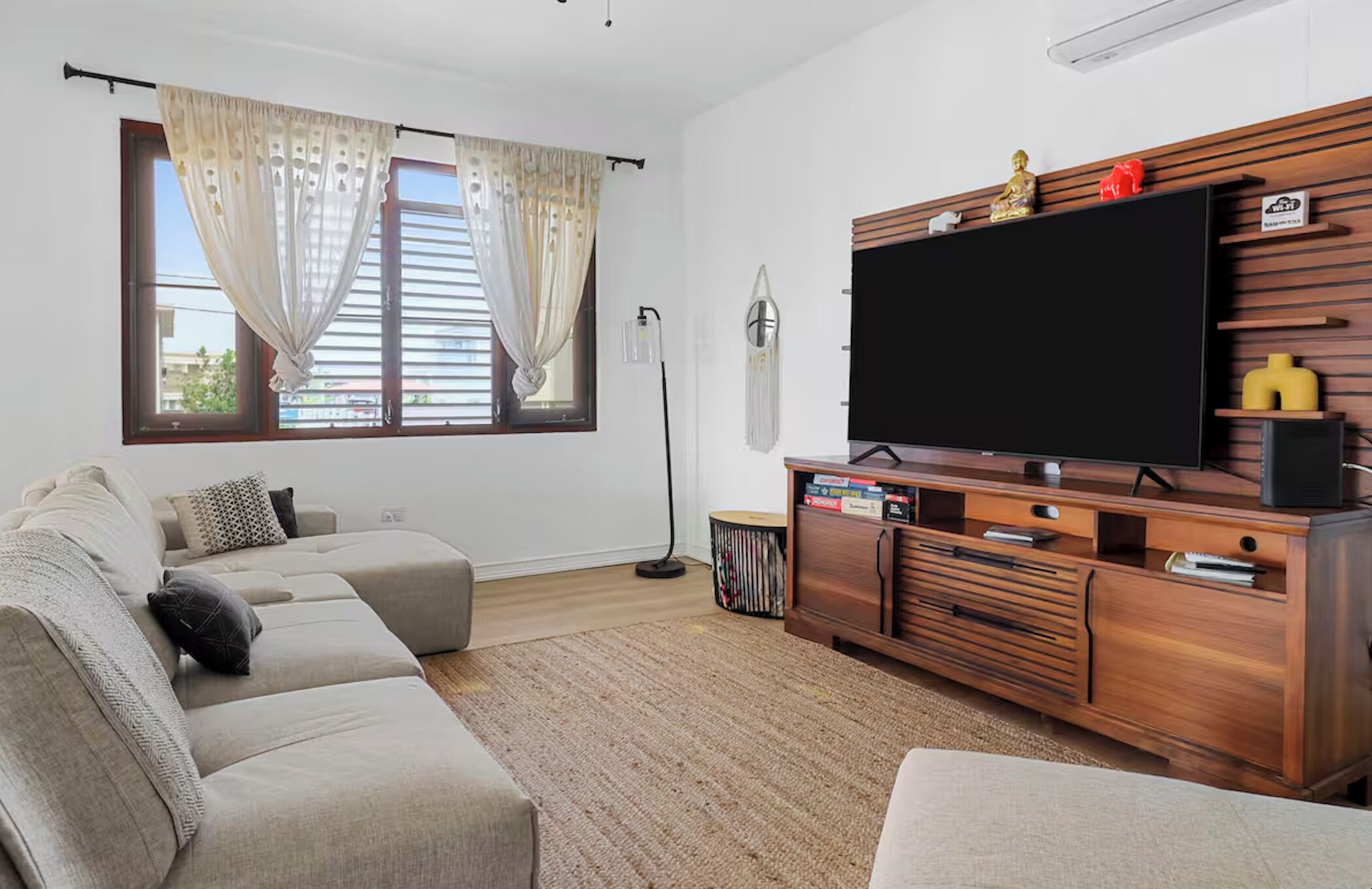 A living room with a beige sofa, a flat-screen TV on a wooden stand, a floor lamp, and a window with curtains.