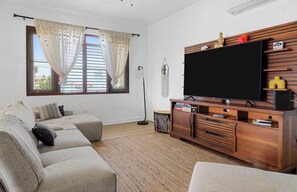 Un salon avec un canapé beige, un téléviseur à écran plat posé sur un meuble en bois, une lampe sur pied et une fenêtre avec des rideaux.