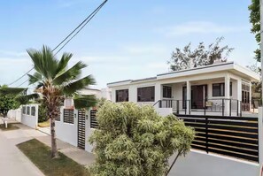 Une maison moderne à deux étages, dotée d’un balcon, entourée d’une clôture blanche et de palmiers.