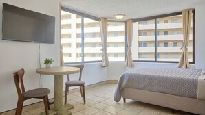 Ein Hotelzimmer mit einem Bett, einem an der Wand befestigten Fernseher, einem kleinen runden Tisch mit zwei Stühlen und großen Fenstern mit Vorhängen.