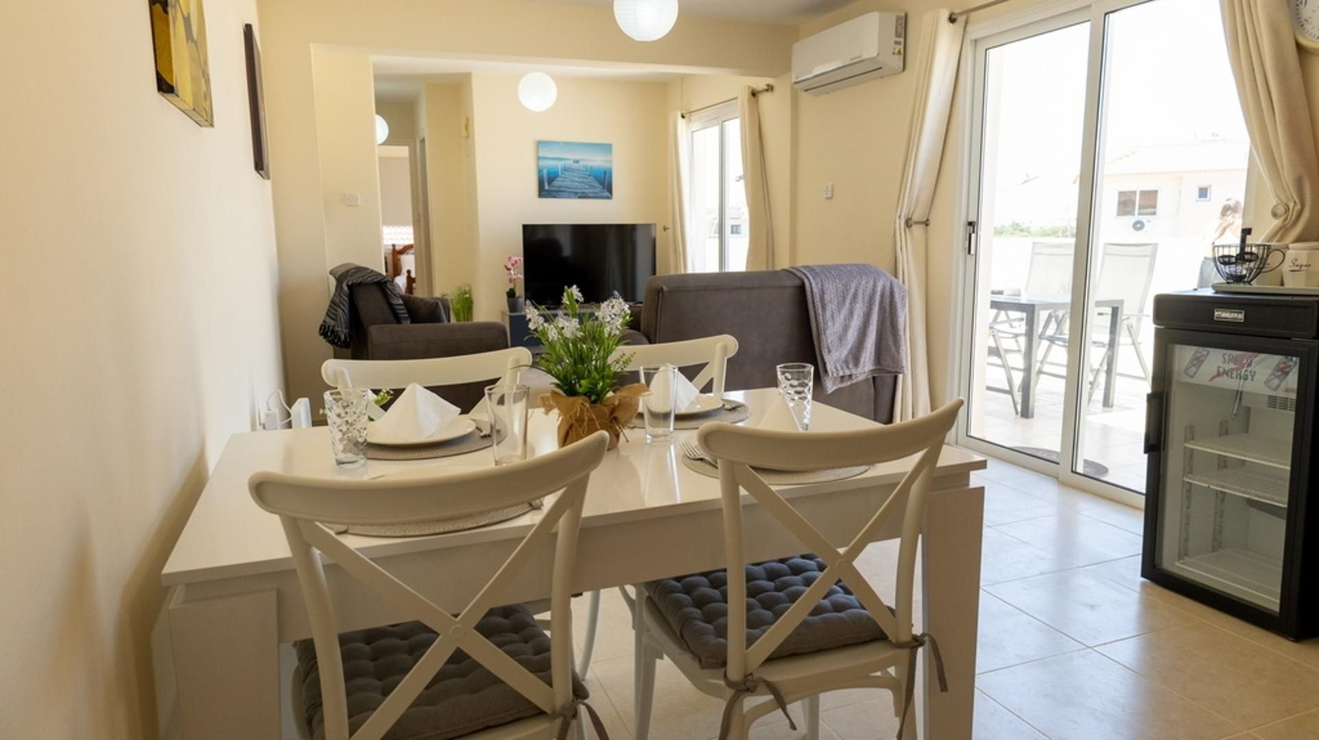 A dining area with a table set for two, a television in the background, and a view of the outside through a glass door.