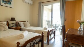 Une chambre d’hôtel avec deux lits, une table de chevet avec une lampe, une chaise en bois, une petite table avec un vase et une fenêtre avec des rideaux.