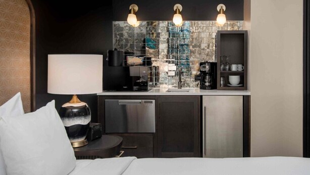 A compact hotel room with a kitchenette, a bed, a bedside table with a lamp, and a TV.