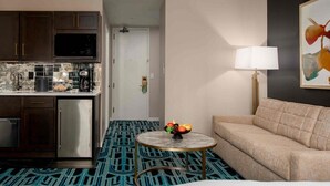 A compact hotel room with a kitchenette, a round coffee table with fruit, a sofa, and a patterned carpet.