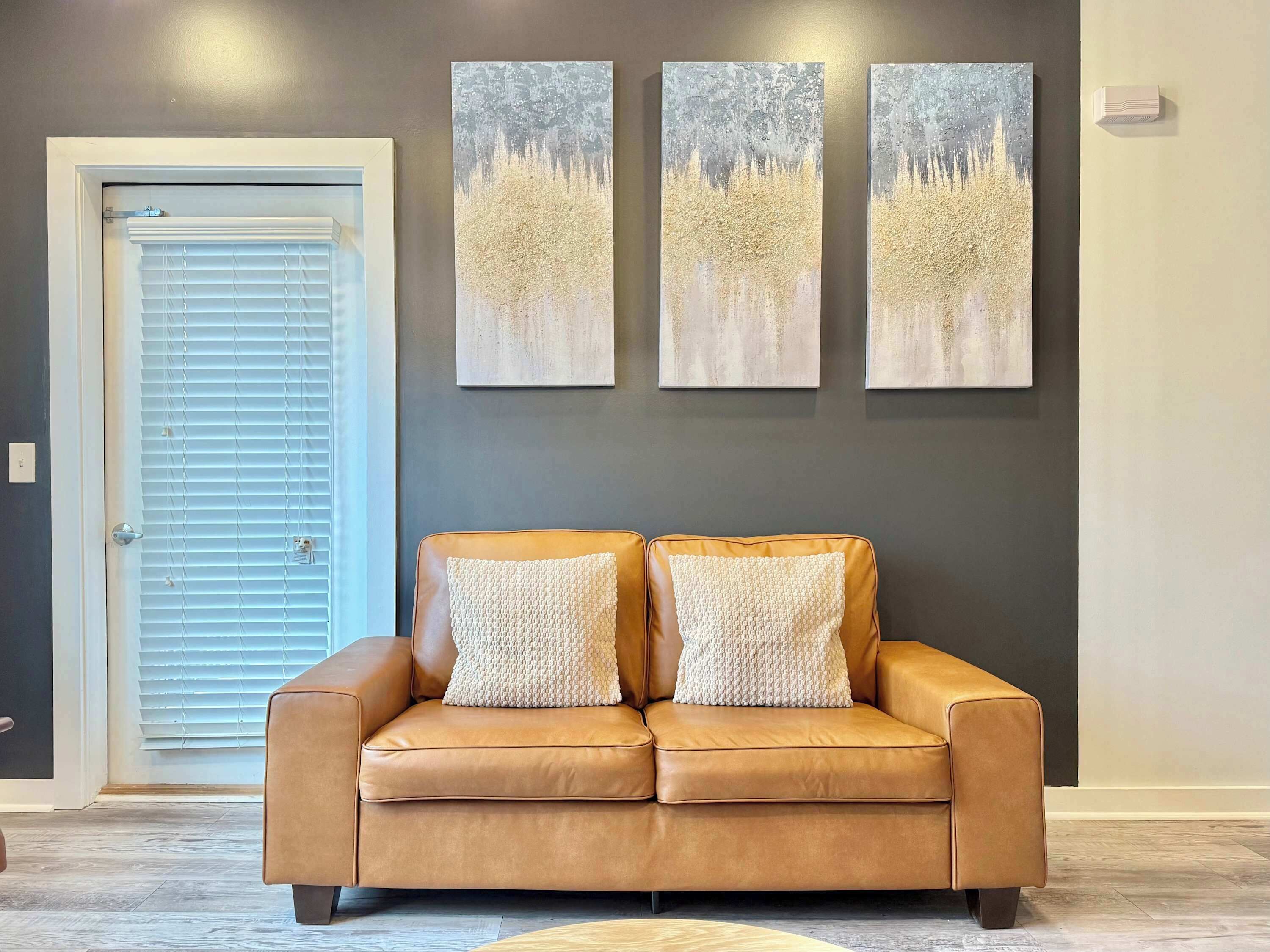 A room with a brown leather sofa, two white textured pillows, and three abstract paintings on the wall.