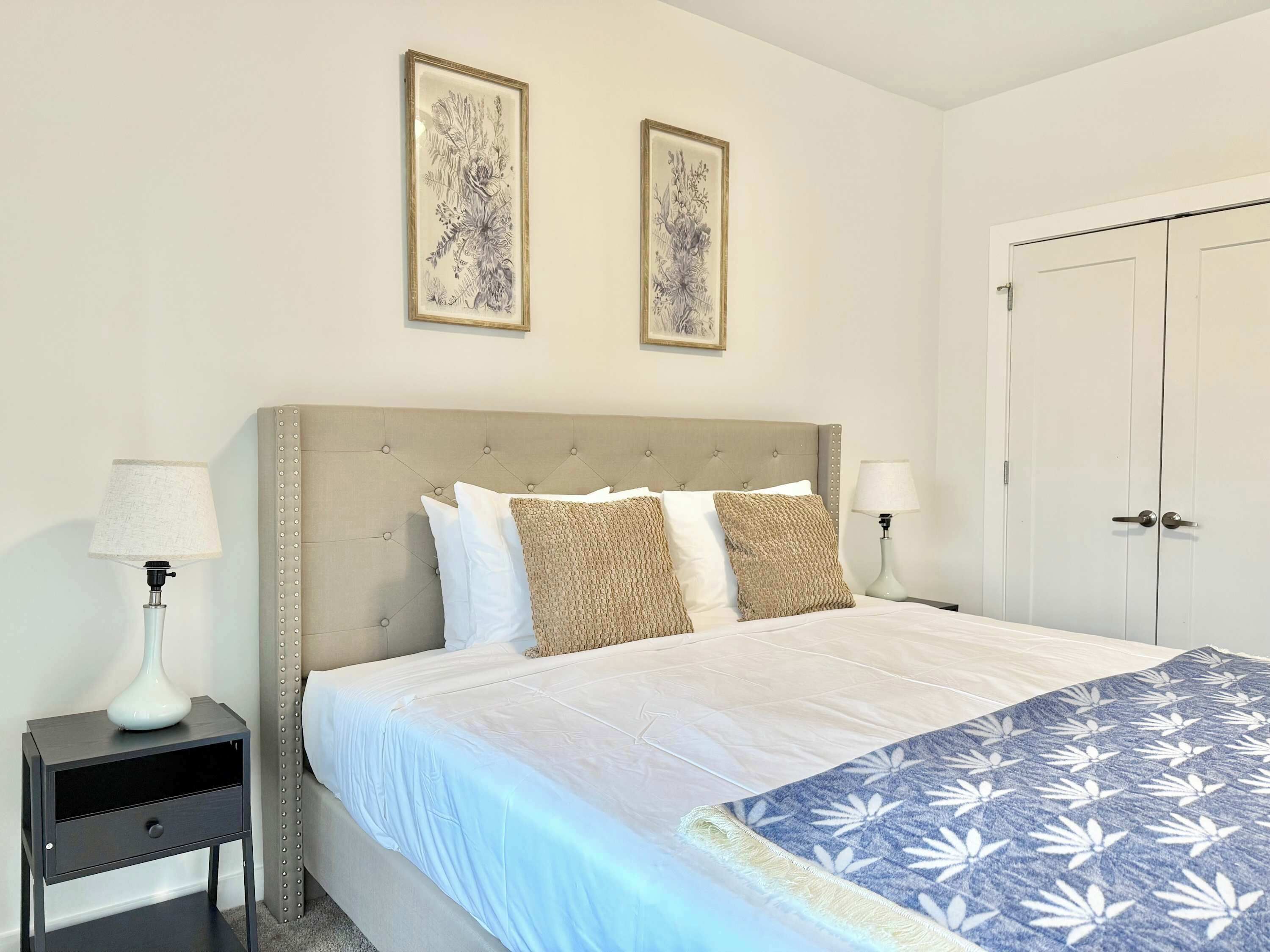 A bedroom with a bed, two bedside tables, a nightstand, a door, and two framed pictures on the wall.