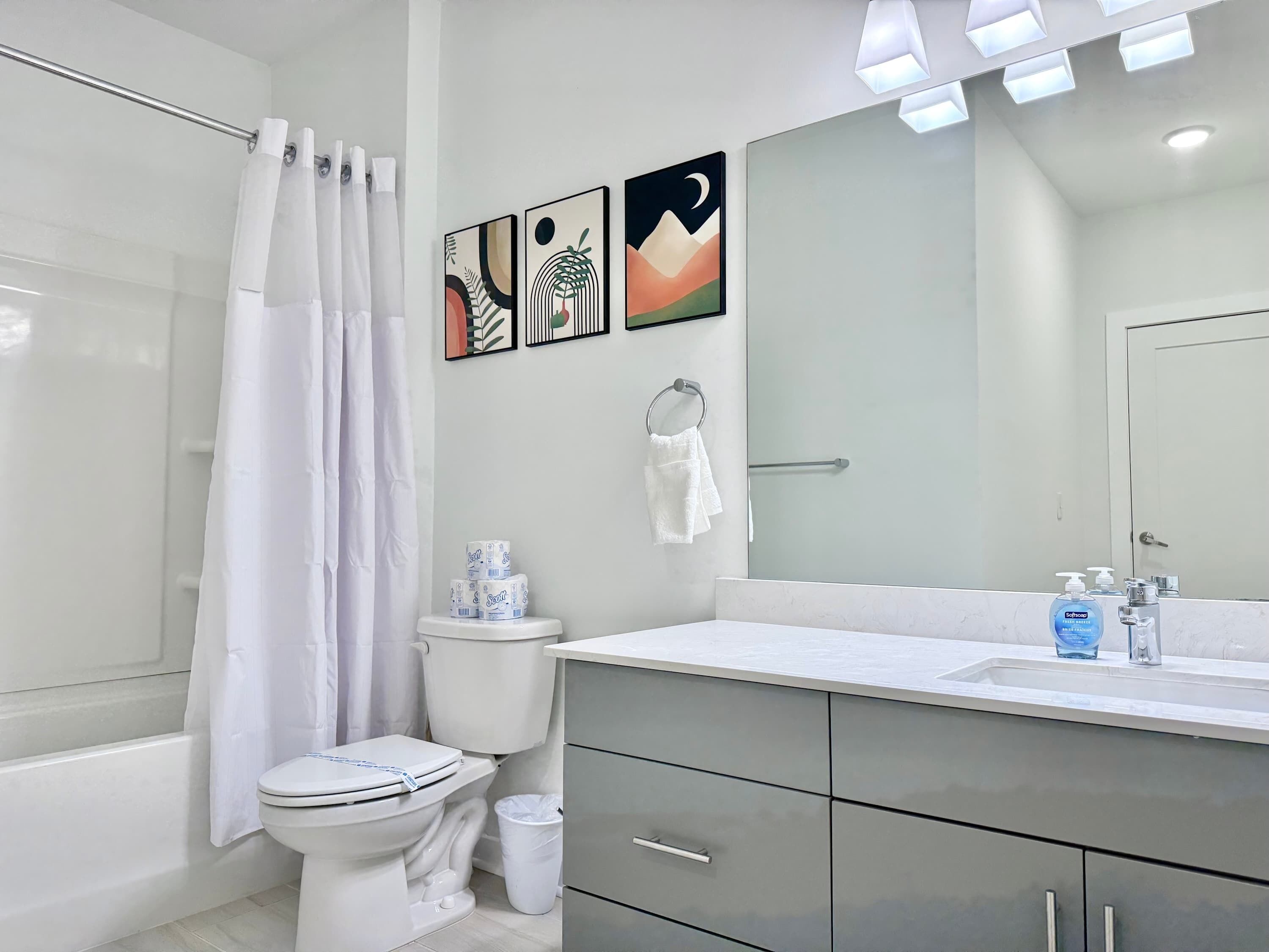 Ein modernes Badezimmer mit Toilette, Bidet und Duschvorhang. An der Wand hängen drei gerahmte Kunstwerke, ein Handtuchhalter und ein Spiegel über dem Waschbecken.