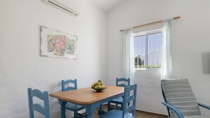 Ein Essbereich mit einem Holztisch, blauen Stühlen, einer Obstschale, einem Fenster mit Vorhängen und einer Wand-Klimaanlage.