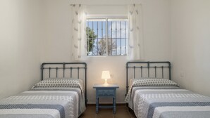 Ein Schlafzimmer mit zwei Betten, einem Nachttisch mit Lampe, einem Fenster mit Gitter und Blick auf Bäume.