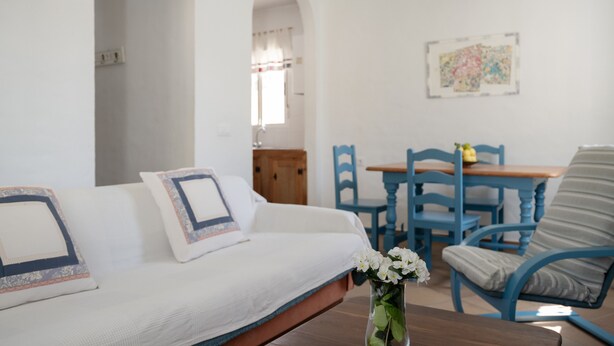 Ein weißes Sofa mit gemusterten Kissen, ein Holztisch, blaue Stühle und eine Vase mit weißen Blumen.
