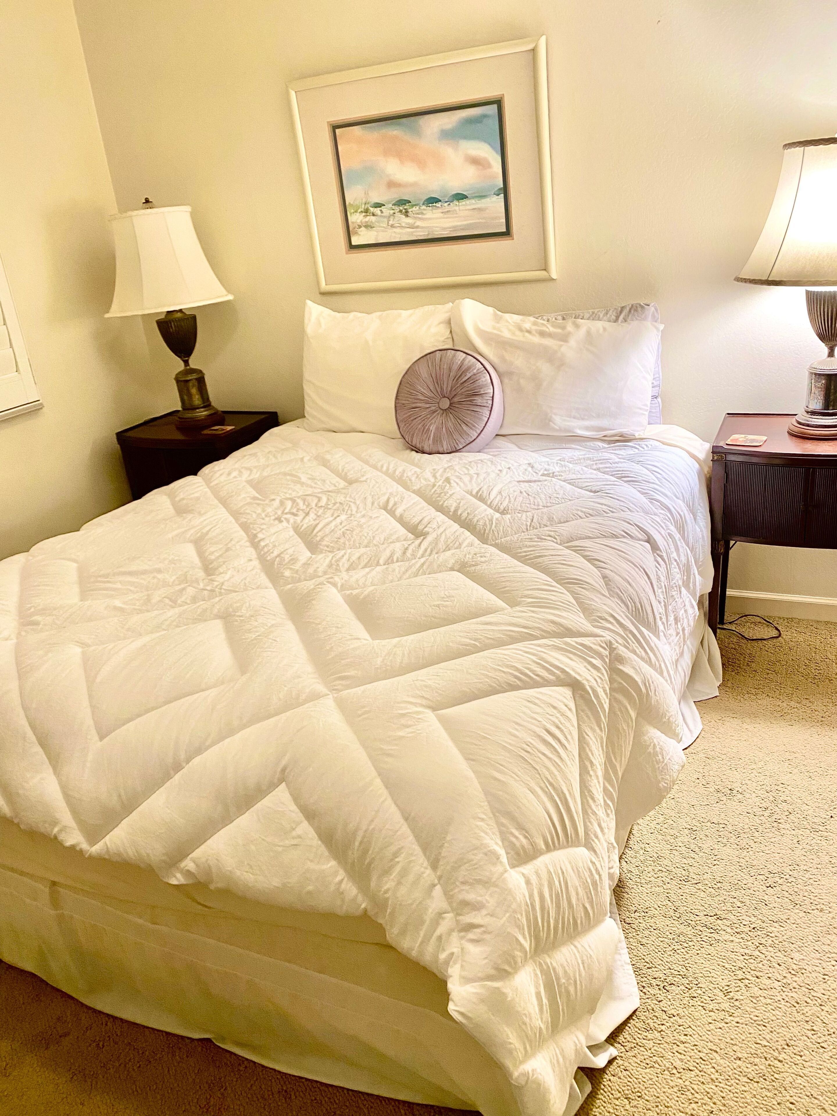 A neatly made bed with a white quilt, a single round pillow, and a framed picture above it.