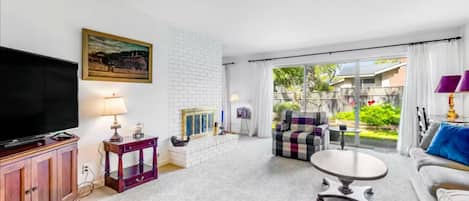 Uma sala de estar com televisão de tela plana, um armário de madeira, uma mesa lateral roxa, uma mesa de centro branca, um sofá e uma janela grande com cortinas.