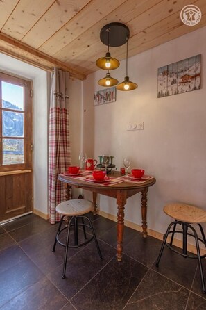 Ein gemütlicher Essbereich mit einem Holztisch, gedeckt für zwei Personen, zwei Barhocker und einem Fenster mit Bergblick.