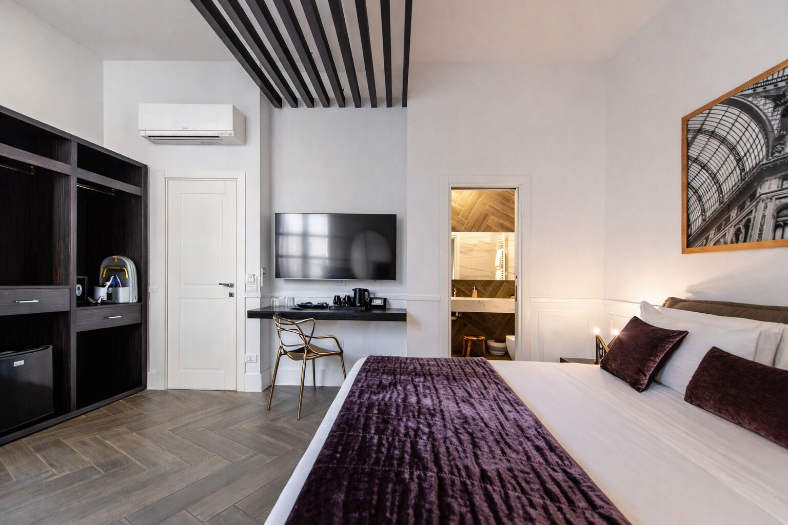 Une chambre d’hôtel moderne équipée d’un lit, d’un bureau, d’une télévision, d’une armoire et d’une salle de bain visible à travers une porte ouverte.