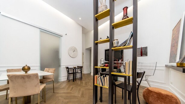 A modern living room with a herringbone wood floor, a round dining table with chairs, a bookshelf, and a large clock on the wall. - Umberto 34 Collection (Naples)