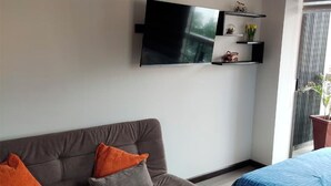 A living room with a grey sofa, orange cushions, a flat-screen TV mounted on the wall, and a bed with a blue bedspread. - Modern studio apartment in downtown area of the city (Cochabamba)