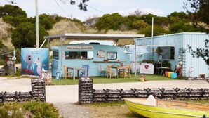 Een klein, lichtblauw gebouw met een bord waarop 'Melshell Oysters' staat. Er is een tafel met stoelen buiten, een klein bootje ervoor en een muurschildering aan de linkerkant.