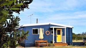 Een laag, eenlaags gebouw met een blauwe gevel, een gele deur en een klein terras met een bank en een parasol.