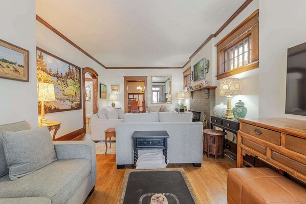 A cozy living room with a grey sofa, a wooden coffee table, and a fireplace. There are framed pictures on the walls and a large mirror above the fireplace.