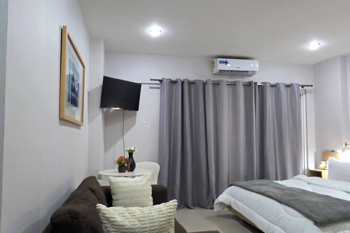 A modern hotel room with a sofa, a bed, a flat-screen TV, and a ceiling-mounted air conditioner.