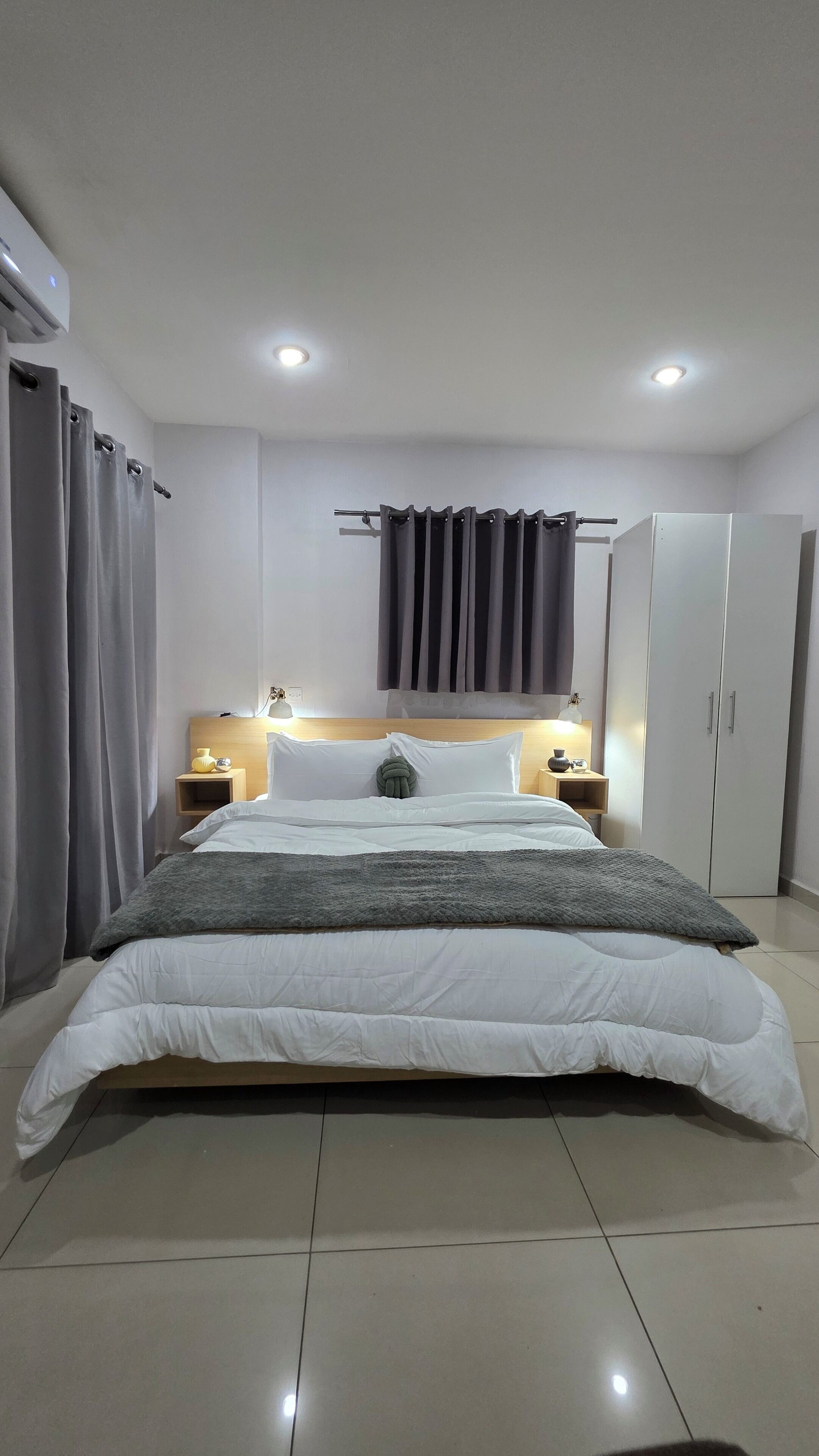 A modern bedroom with a large bed, a wooden headboard, two bedside tables with lamps, a wardrobe, and grey curtains.